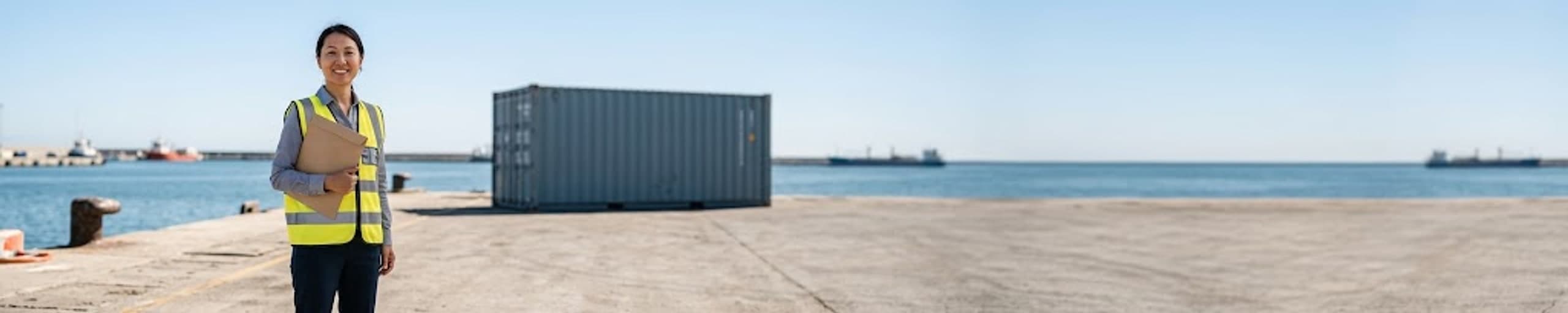 Responsable export contrôlant les documents d'expédition devant un conteneur en cours de chargement d'engins de chantier, terminal portuaire à l'arrière-plan