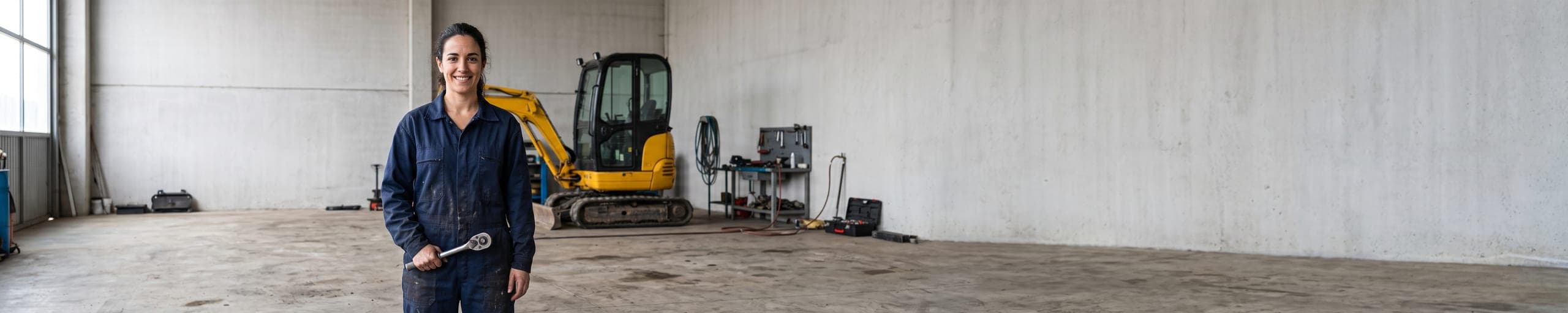 Mécanicienne effectuant l'entretien du compartiment moteur ouvert d'une mini-pelle jaune dans un atelier de réparation, autres engins visibles à l'arrière-plan